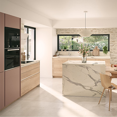 une grande cuisine claire, avec un grand îlot central. L’îlot, tout en longueur, est divisé en une moitié plan de travail en marbre, et une autre moitié étant une table en bois claire autour de laquelle sont installées 4 chaises. Des assiettes sont sur cette table pour illustrer que l’on peut s’y attabler pour un repas.