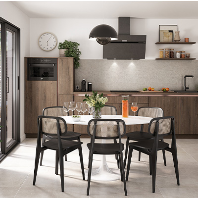 la cuisine Luna d’ixina une cuisine de style industriel, aux murs blancs avec placards en bois foncé, aux accents en métal noir. Au premier plan, une table et des chaises noires avec empiècement en rotin clair. La décoration est minimaliste, avec quelques plantes vertes et des étagères ouvertes sur lesquelles figurent des bocaux en verre.
